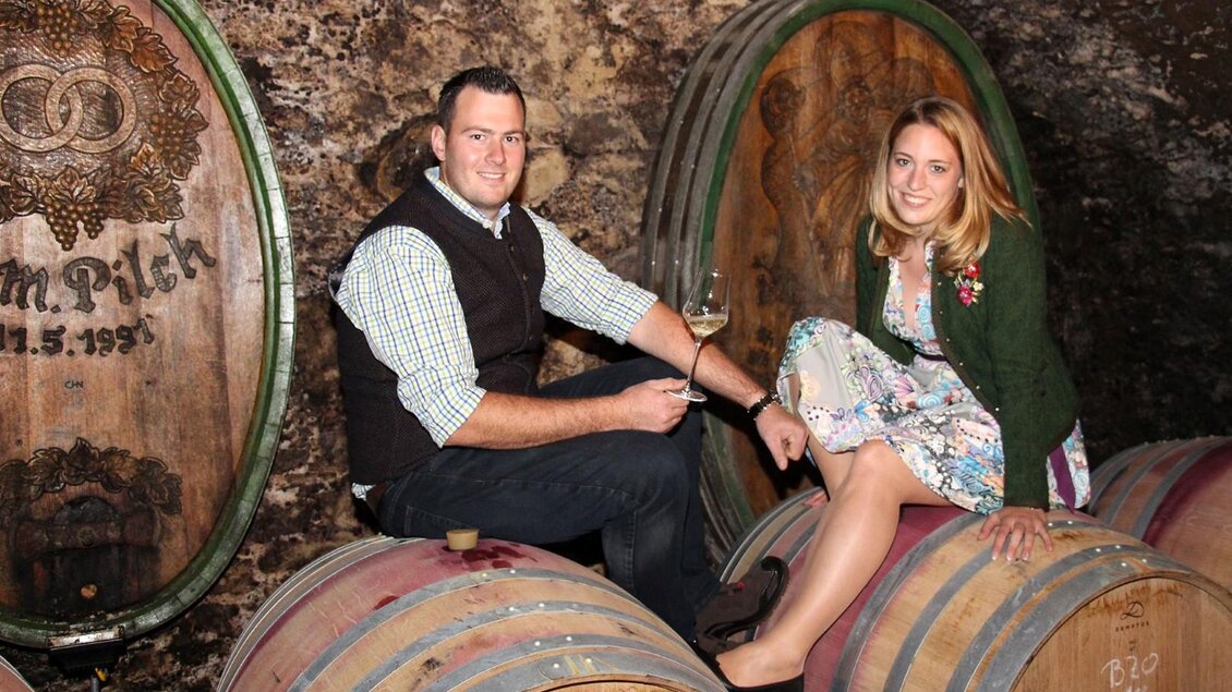 Ein Paar sitzt auf Weinfässern in einem Weinkeller. Im Hintergrund sind Holzfässer mit Dekorationen zu sehen. | © Weingut Pilch | Fam. Pilch