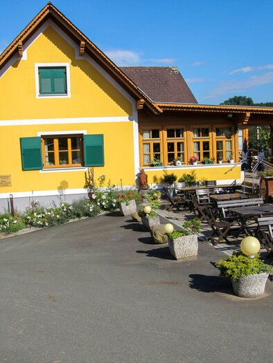 A picturesque yellow house with green shutters. Outside, there are cozy seating areas and blooming plants. | © Buschenschank Rank