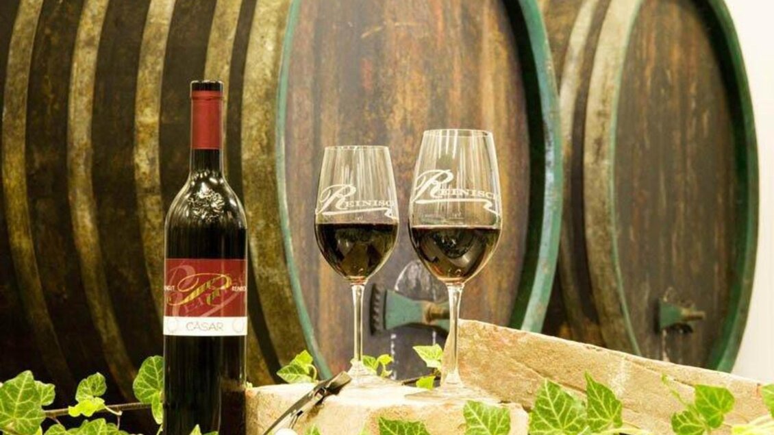 Eine Flasche Rotwein steht neben zwei Weingläsern. Im Hintergrund sind große, traditionelle Weinfässer zu sehen. | © Weingut Reinisch