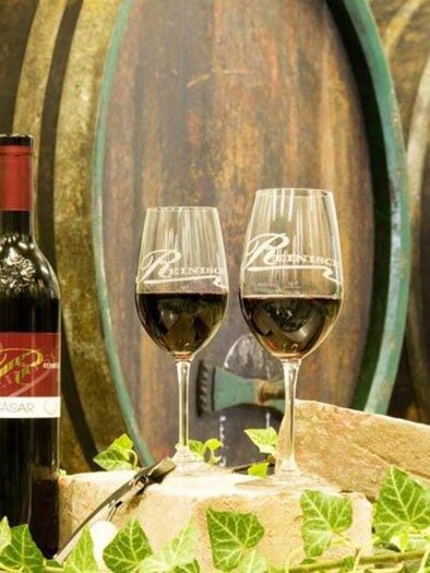 A bottle of red wine stands next to two wine glasses. In the background, large, traditional wine barrels can be seen. | © Weingut Reinisch