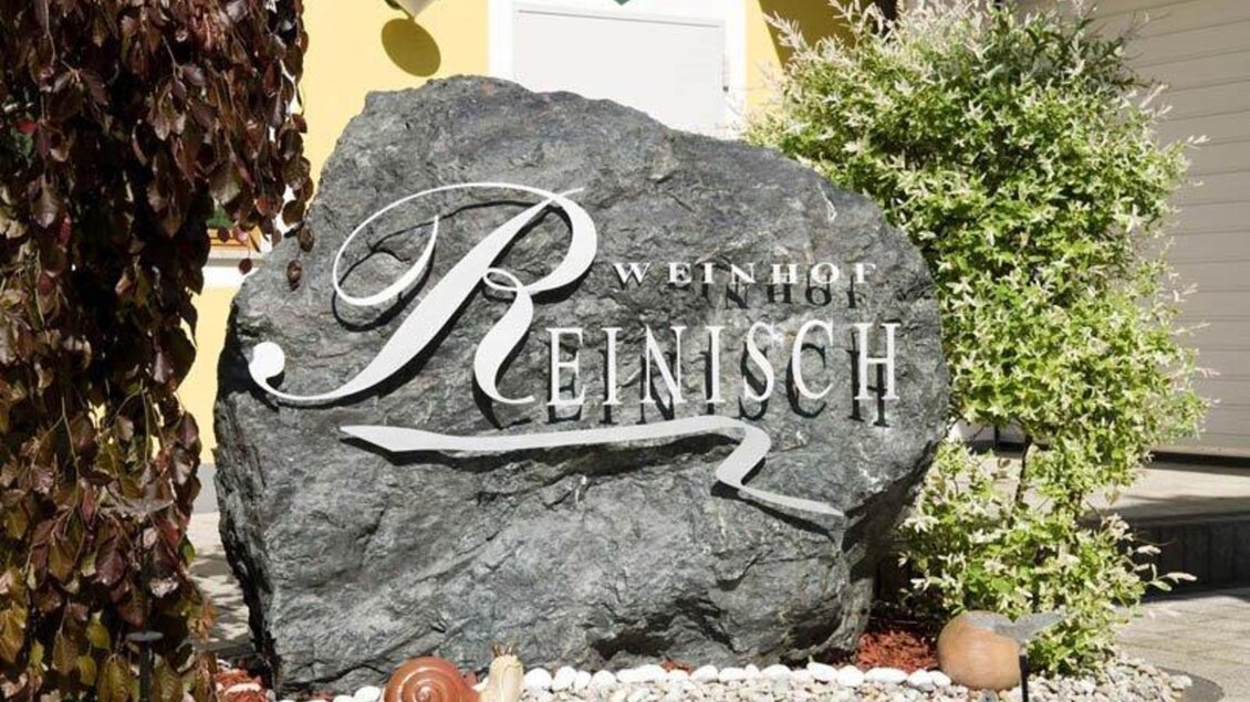 Ein dekorativer Felsen mit der Aufschrift „Weinhof Reinisch“ steht im Garten. Umgeben von Pflanzen und Kies schafft er eine einladende Atmosphäre. | © Weingut Reinisch