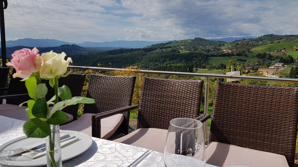Ein eleganter Tisch mit Blumen und Glaswaren, umgeben von einem schönen Blick auf grüne Hügel und eine hügelige Landschaft. Der Himmel ist leicht bewölkt, was eine entspannte Atmosphäre schafft. | © Weingut Reiterer