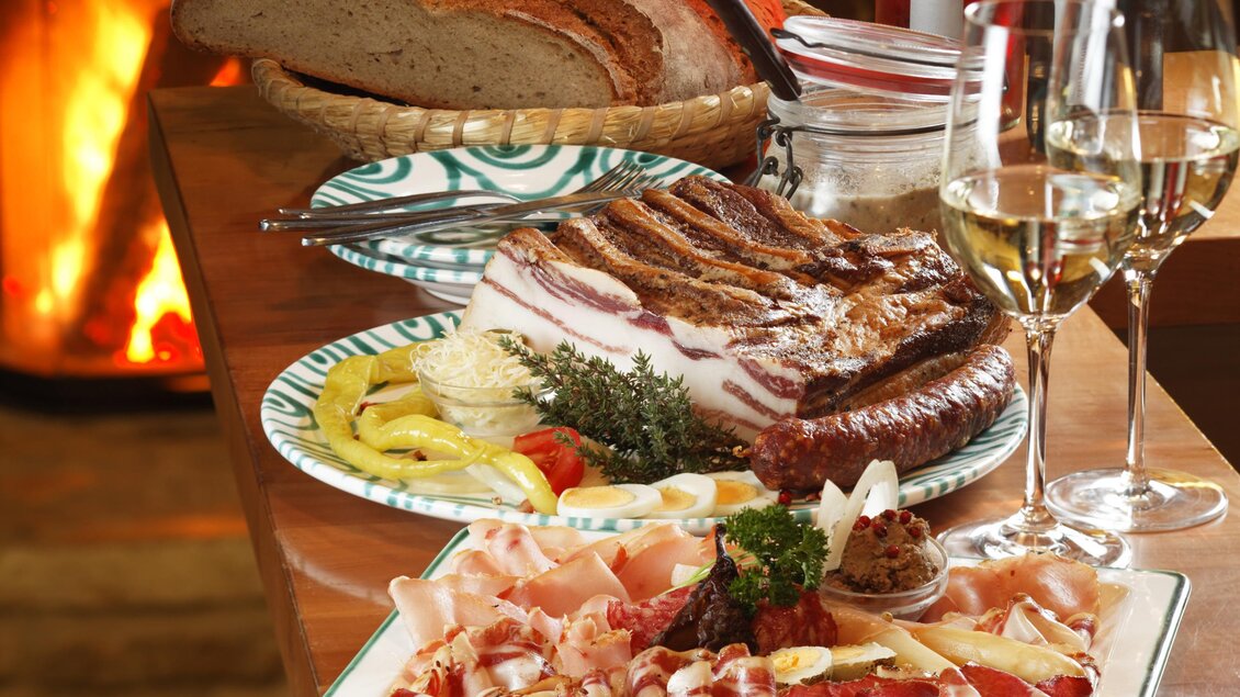 Ein rustikaler Tisch mit verschiedenen Fleischspezialitäten, Brot und köstlichen Beilagen. Im Hintergrund brennt ein Kamin, und auf dem Tisch stehen Weingläser und Flaschen. | © Buschenschank Retter Kneissl