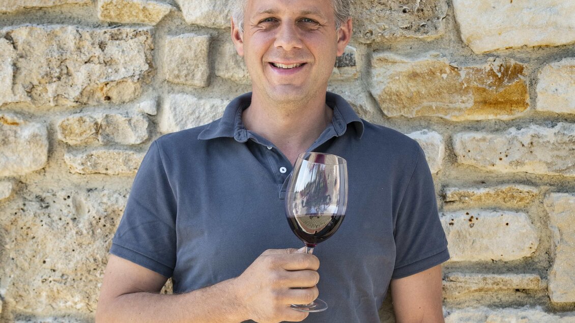 Ein Mann steht vor einer steinernen Wand und hält ein Glas Rotwein in der Hand. Er lächelt freundlich in die Kamera. | © Oststeiermark Tourismus