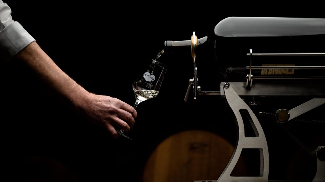 Ein elegantes Gerät zum Einschenken von Wein wird verwendet. Ein Glas wird mit einem klaren Getränk gefüllt, während der Hintergrund dunkel ist. | © Weingut | Buschenschank Strauss