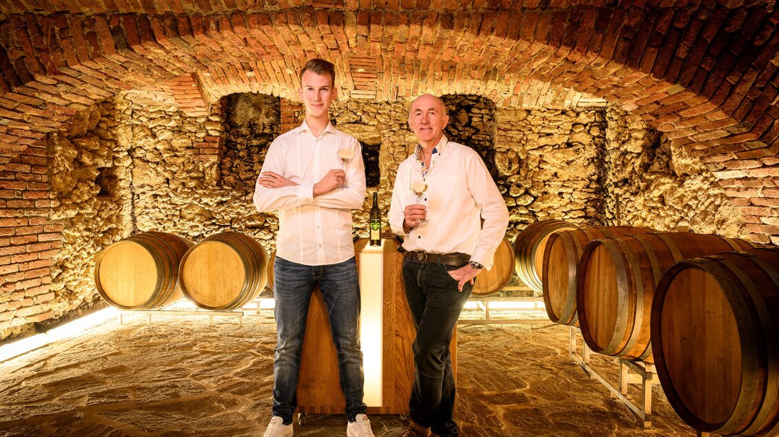 Zwei Männer stehen in einem Weinkeller zwischen Holzfässern. Die Wände sind aus Stein und das Licht schafft eine gemütliche Atmosphäre. | © Weingut | Buschenschank Strauss
