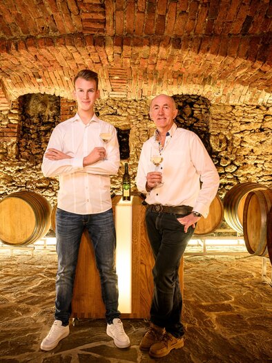 Two men are standing in a wine cellar among wooden barrels. The walls are made of stone and the light creates a cozy atmosphere. | © Weingut | Buschenschank Strauss