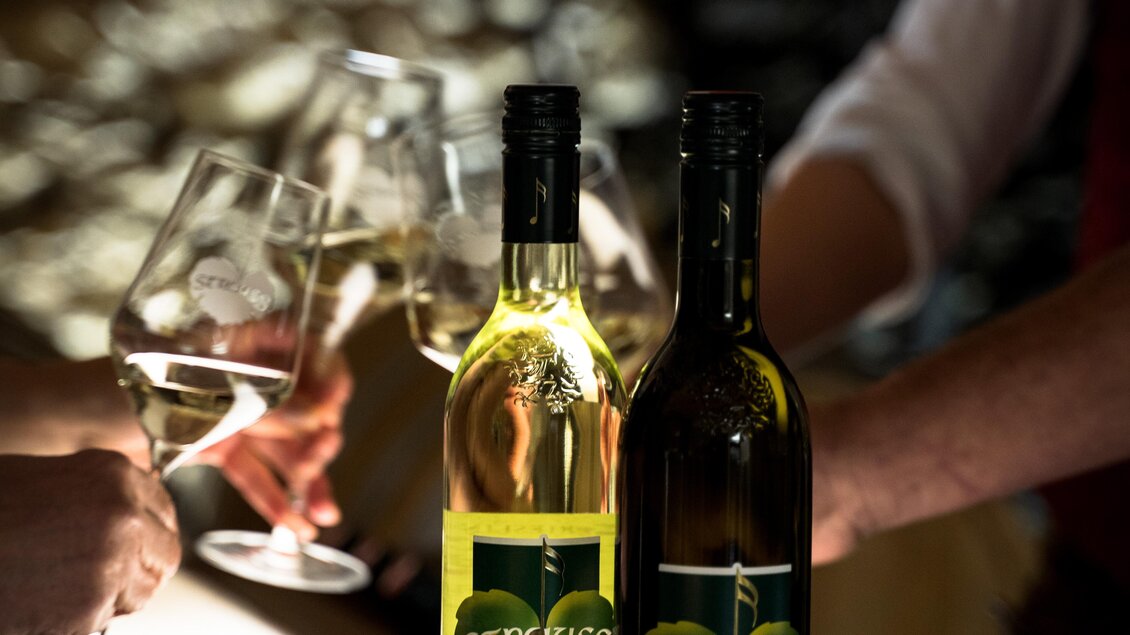 Zwei Flaschen Wein stehen auf einem Tisch, eine davon ist ein Weißwein und die andere ein Rotwein. Im Hintergrund sind Glasbecher und Hände zu sehen, die anstoßen. | © Weingut | Buschenschank Strauss