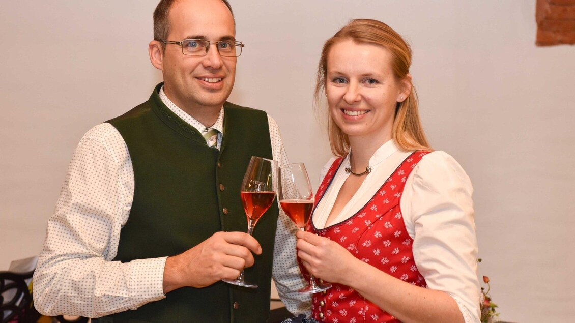 Ein festlich gekleidetes Paar stößt mit Gläsern an. Der Mann trägt eine traditionelle Tracht, die Frau ein Dirndl. | © Weinhof Tropper