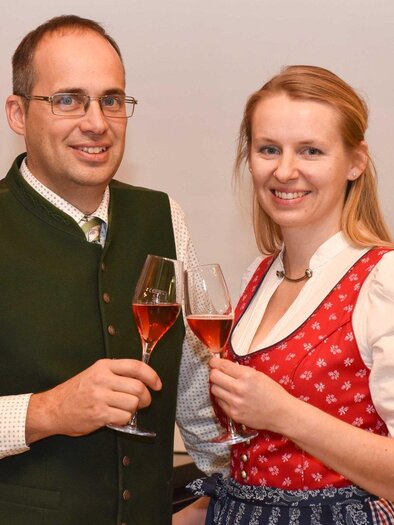 A festively dressed couple toasts with glasses. The man is wearing traditional attire, and the woman is in a dirndl. | © Weinhof Tropper