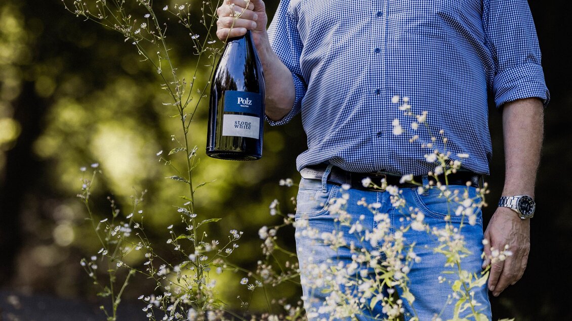 Ein Mann mit lockigem Haar steht in einer grünen Umgebung und hält eine Flasche Sekt in der Hand. Er trägt ein blaues Hemd und eine entspannte Ausstrahlung. | © Weingut Walter Polz