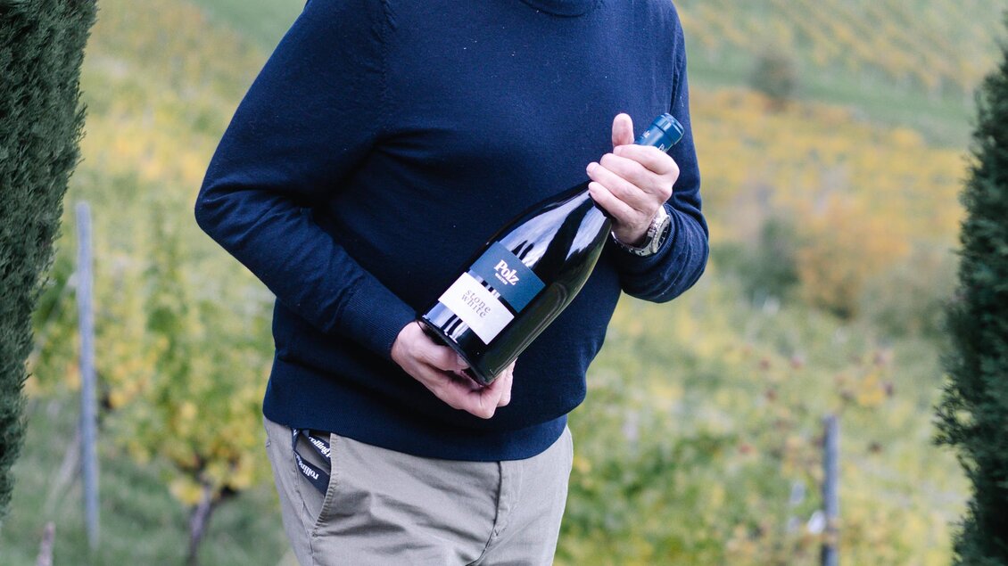 Ein Mann steht in einem Weinberg und hält eine Flasche Wein in der Hand. Im Hintergrund sind grüne Weinreben und sanfte Hügel zu sehen. | © Renate Polz