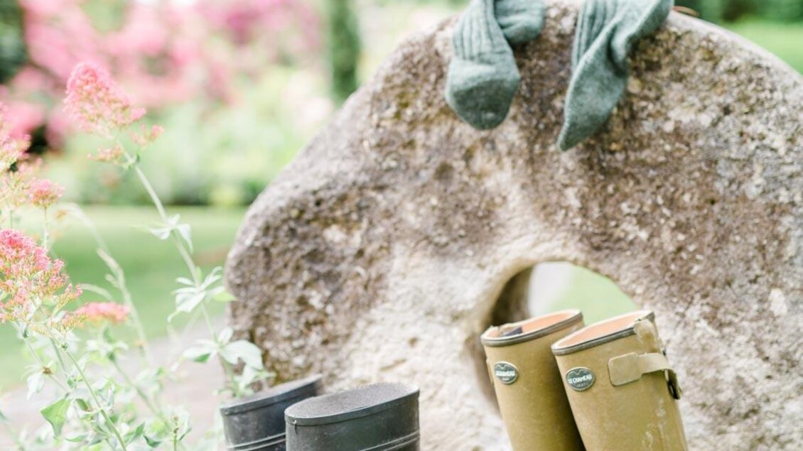 Gummistiefel stehen neben einem großen Stein im Garten. Im Hintergrund sind Blumen und Bäume zu sehen. | © Renate Polz