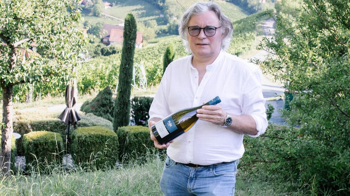 Ein Mann steht in einem Weinberg und hält eine Flasche Sekt in der Hand. Im Hintergrund sind Hügel und Bäume zu sehen. | © Renate Polz