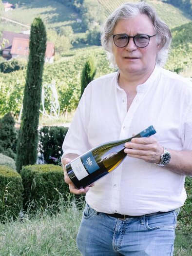 A man is standing in a vineyard holding a bottle of sparkling wine in his hand. In the background, hills and trees can be seen. | © Renate Polz