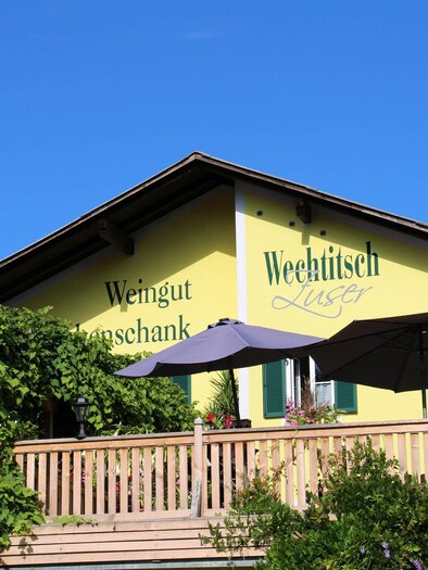 A yellow building with a terrace area, shaded by a large parasol. It bears the inscription "Weingut Lusser" and is surrounded by green plants. | © Weingut Wechtitsch