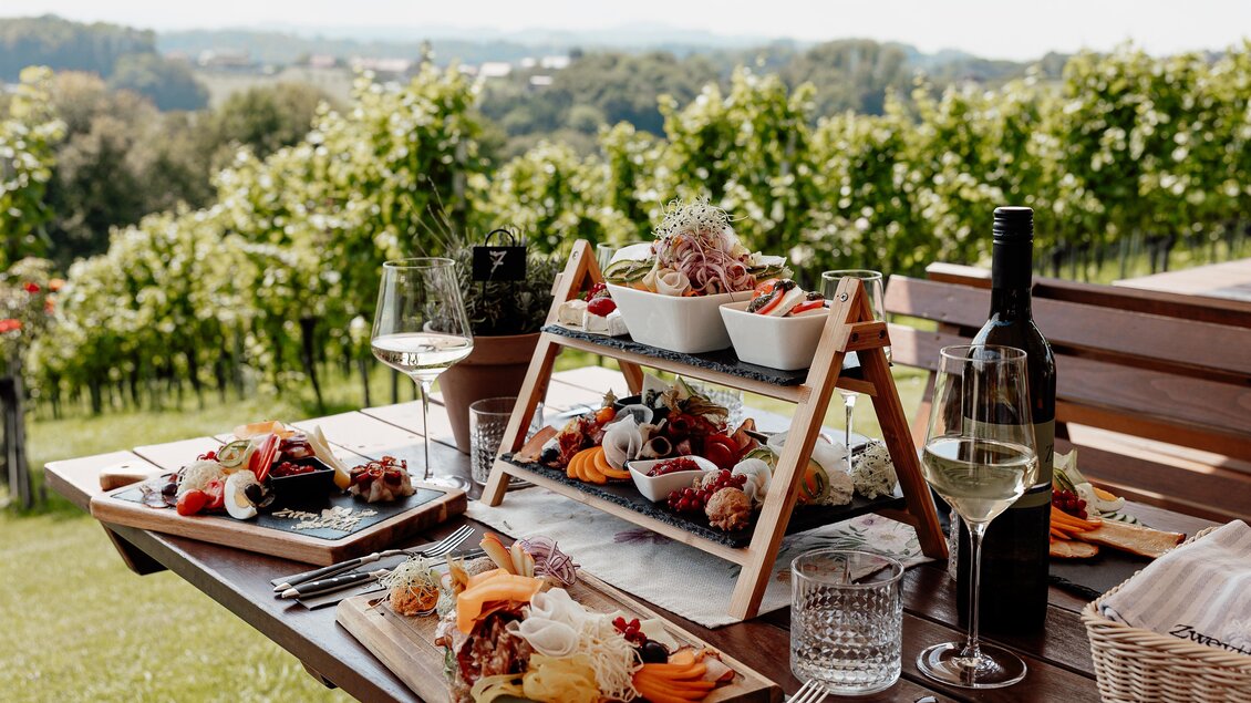 Ein schöner gedeckter Tisch im Freien mit einer Vielzahl an leckeren Speisen und einer Flasche Wein. Im Hintergrund sind Weinberge und eine malerische Landschaft zu sehen. | © Katja Koller