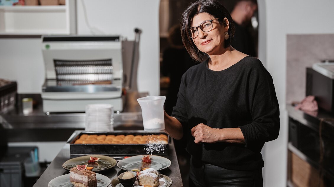 Waltraud steht in Küche bei verschiedenen Desserts, einschließlich Kuchen. Sie lächelt und hält einen Messbecher in der Hand. | © Michaela Lorber