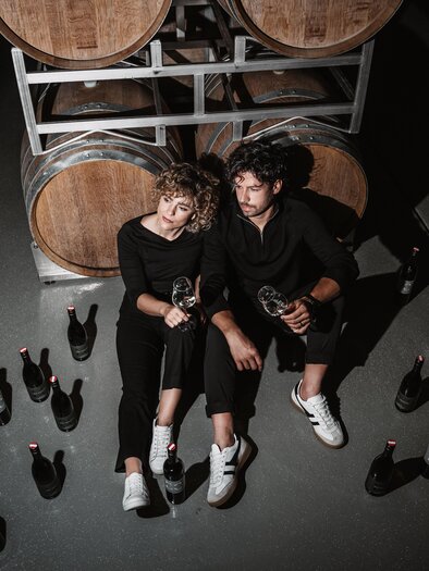A couple is sitting casually on the floor of a wine cellar, holding wine glasses in their hands. Surrounding them are numerous wine bottles and wooden barrels in the background. | © Michaela Lorber