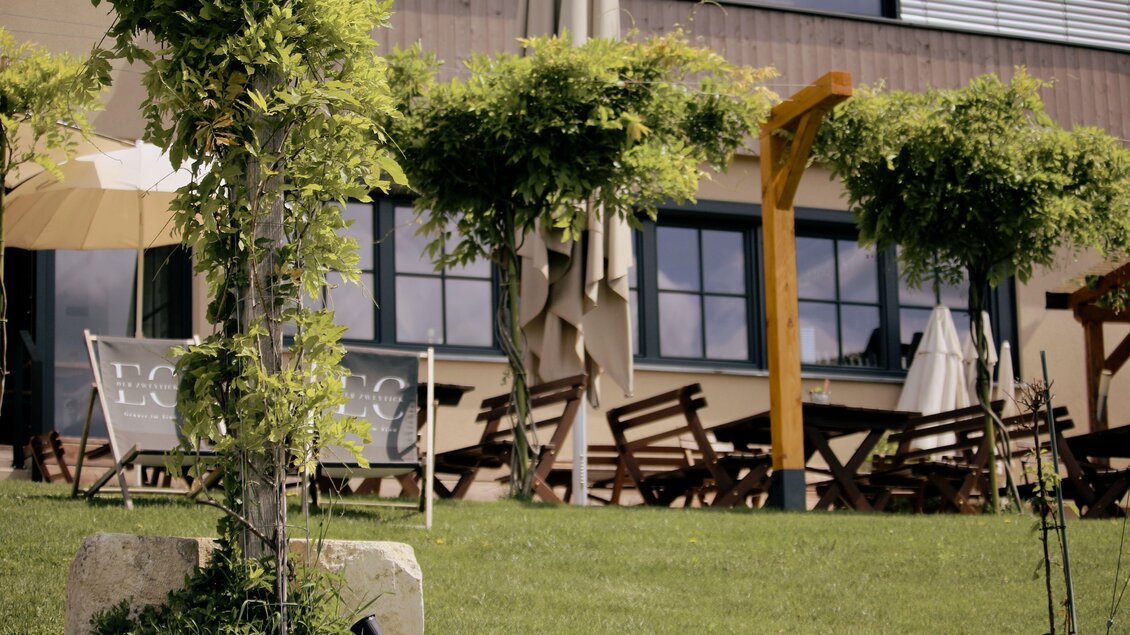 Ein einladender Außenbereich eines Restaurants mit grünen Bäumen und gemütlichen Sitzgelegenheiten. Sonnenschirme sorgen für Schatten an einem schönen Tag. | © Elena Zweytick