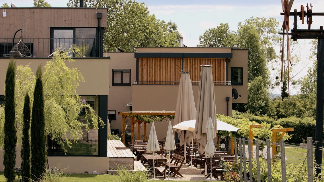 Ein modernes Gebäude mit einem Gartenbereich. Tische und Sonnenschirme sind für Gäste im Freien hergerichtet. | © Elena Zweytick