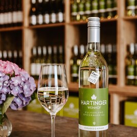 Eine Flasche Hartinger Weißwein steht neben einem Weinglas auf einem Tisch. Im Hintergrund sind Regale mit weiteren Weinflaschen und einer blühenden Pflanze zu sehen. | © Weinhof Hartinger