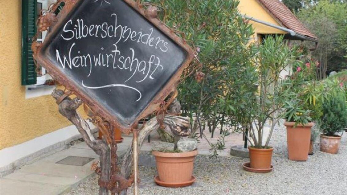 Ein dekoratives Schild aus Holz mit der Aufschrift „Silberschneider's Weinwirtschaft“ steht auf einem Kiesplatz. Im Hintergrund sind bepflanzte Töpfe und ein landwirtschaftliches Gebäude zu sehen. | © Weinhof Silberschneider