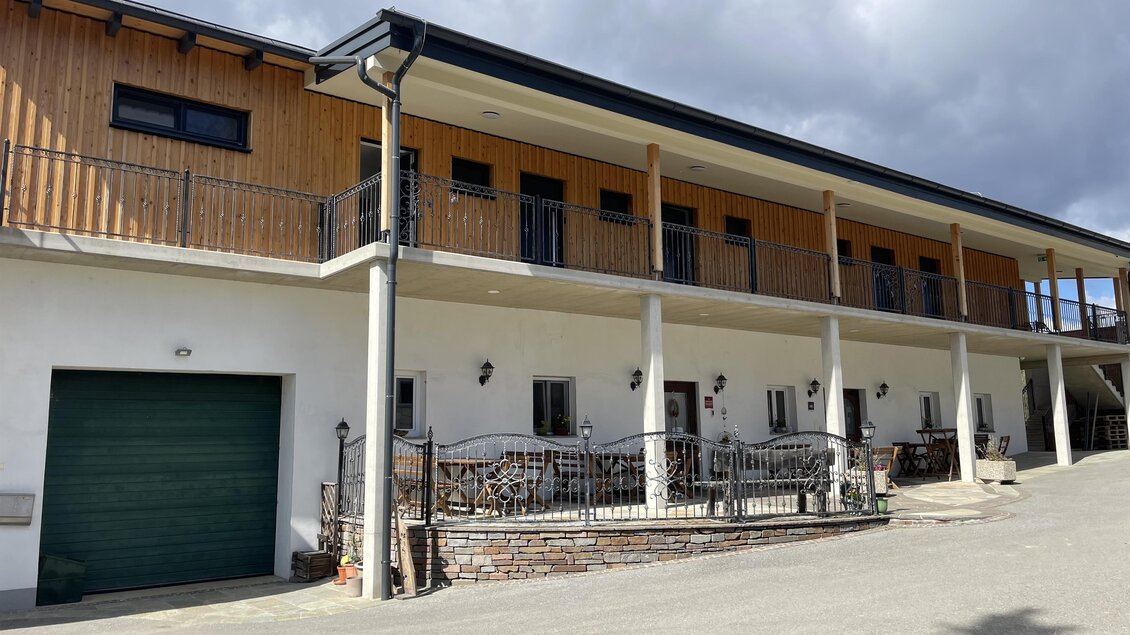 Ein modernes Gebäude mit Holzverkleidung und einer Veranda. Vor dem Haus befinden sich einige Sitzgelegenheiten und ein Garagentor. | © Weinhof Tropper