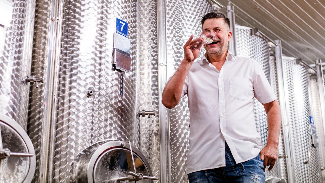 Ein Mann mit einem Glas in der Hand steht in einer Weinkellerei. Im Hintergrund sind große, silberne Gärtanks zu sehen. | © Florfotografie