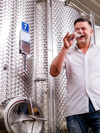 Ein Mann mit einem Glas in der Hand steht in einer Weinkellerei. Im Hintergrund sind große, silberne Gärtanks zu sehen. | © Florfotografie