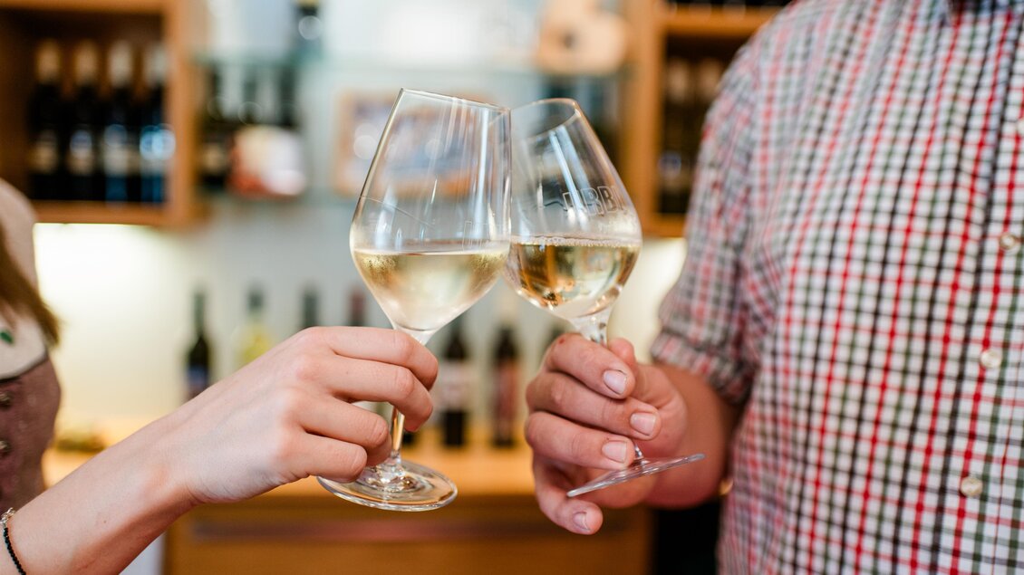 Zwei Gläser Weißwein werden angestoßen. Im Hintergrund sind Weinflaschen zu sehen. | © Florfotografie