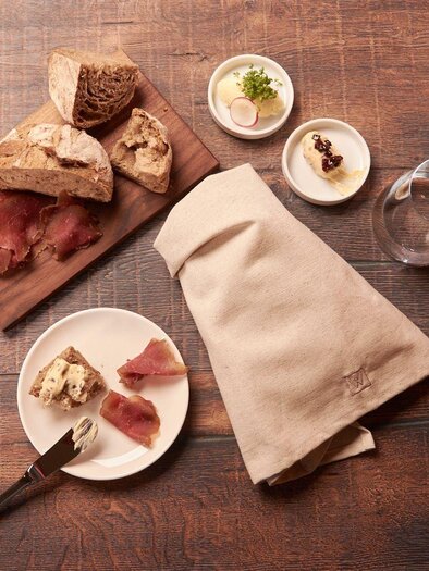 Ein rustikaler Tisch mit verschiedenen Brotsorten und Aufschnitt. Neben einem Teller mit Snacks und einem Glas Wasser. | © Weinkehr