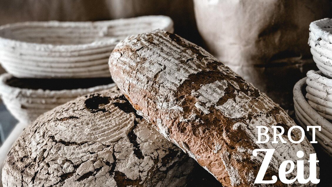 Ein herzhaftes Weizenvollkornbrot liegt auf einer rustikalen Holzoberfläche. Im Hintergrund sind einige Körbe sichtbar, die das handwerkliche Ambiente unterstreichen. | © Fam. Paar