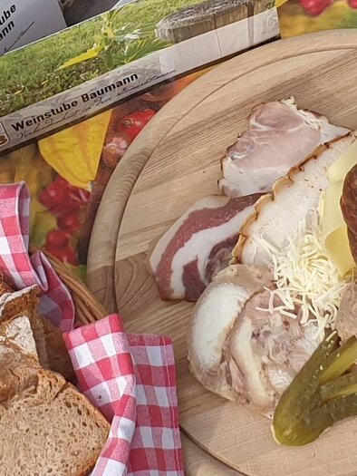 Ein rustikales Brett mit herzhaftem Essen, darunter verschiedene Fleischsorten, Sauerkraut und frisches Gemüse. Neben dem Brett liegt ein Laib Brot mit einer karierten Serviette. | © Weinstube Baumann