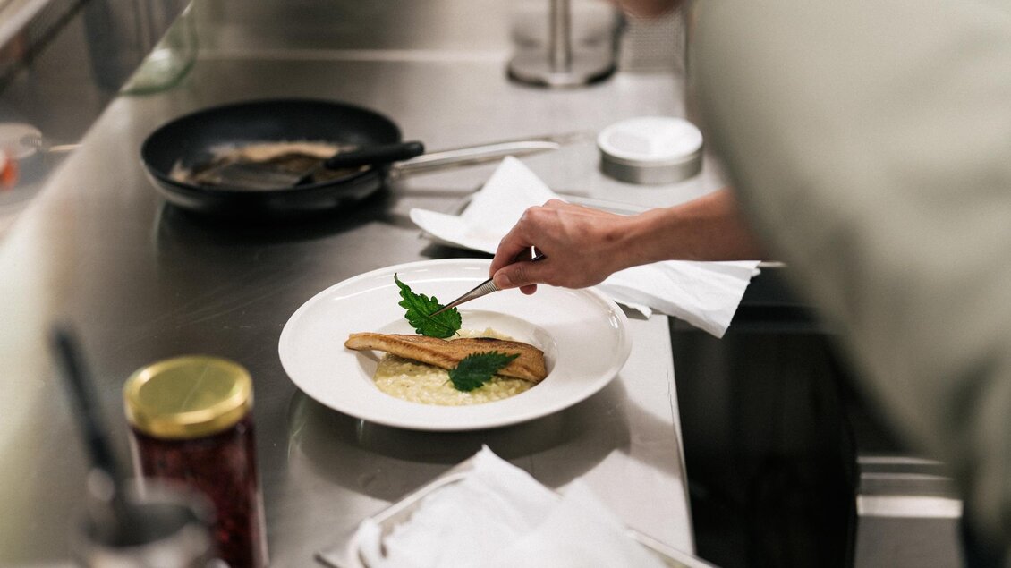 Ein Koch richtet ein Gericht mit Fisch an und dekoriert es mit frischen Kräutern. Im Hintergrund sind weitere Kochutensilien und Lebensmittel sichtbar. | © Weizerhütte