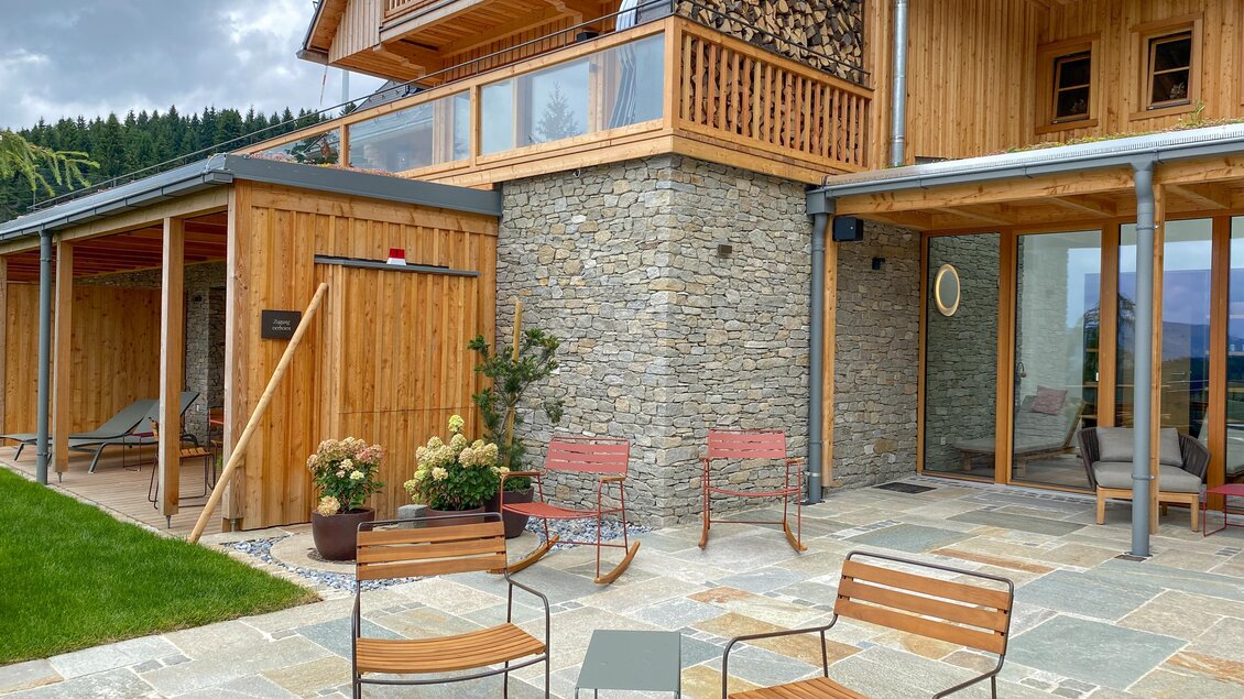 Ein modernes Holzhaus mit Steinelementen und großen Fenstern. Im Vordergrund stehen zwei Stühle auf einer Terrasse, umgeben von einem gepflegten Garten. | © Oststeiermark Tourismus
