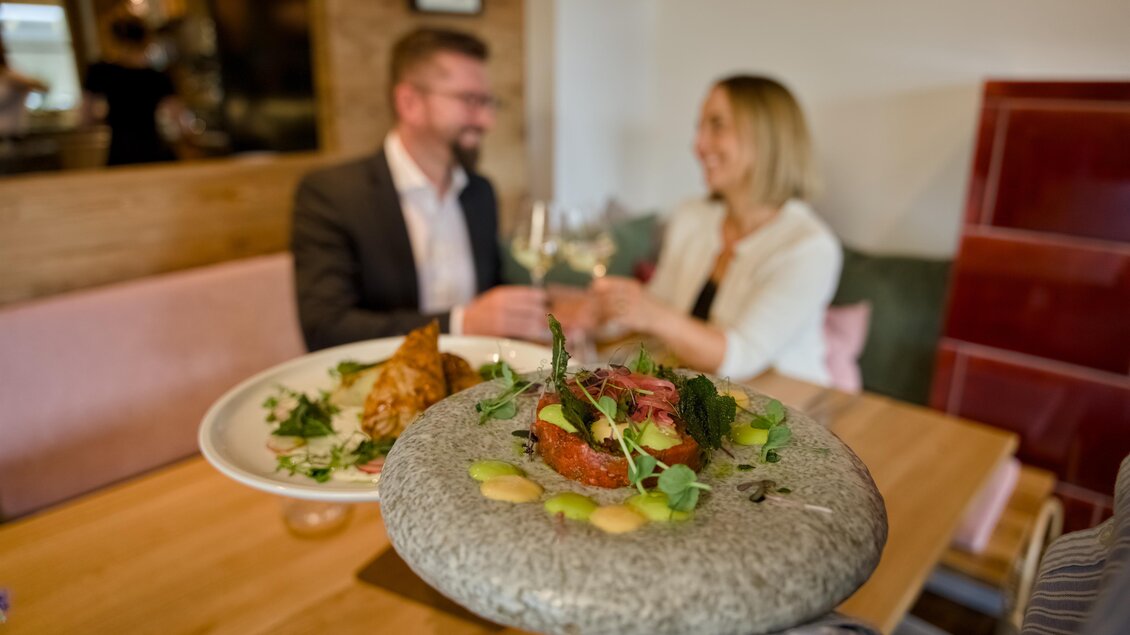 Ein stilvolles Restaurant mit einem eleganten Gericht auf einem Steinplateau. Im Hintergrund stoßen ein Mann und eine Frau mit Gläsern an. | © Oststeiermark Tourismus