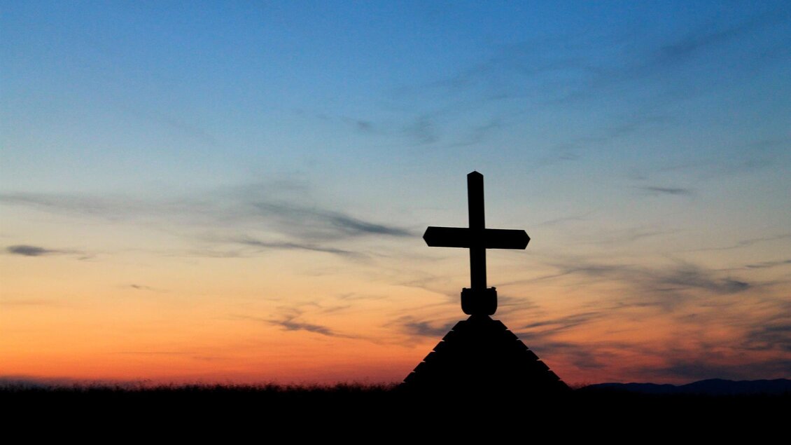 Ein Kreuz auf einem Hügel silhouettiert gegen einen bunten Sonnenuntergang. Der Himmel zeigt sanfte Farben von Blau und Orange. | © Tourismusverband Oststeiermark