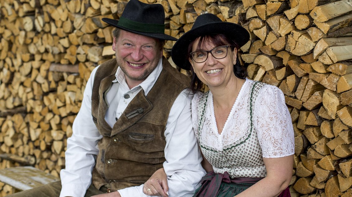 Ein glückliches Paar sitzt vor einem Holzstapel. Beide tragen traditionelle Kleidung und lächeln freundlich in die Kamera. | © Gasthof Orthofer