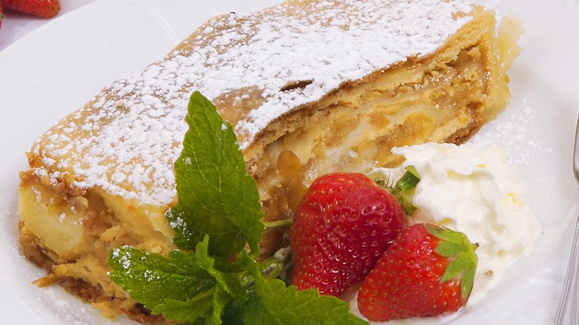 Ein köstlicher Apfelstrudel mit Puderzucker, serviert mit Sahne und frischen Erdbeeren. Das Gericht liegt dekorativ auf einem weißen Teller. | © Gasthof Orthofer