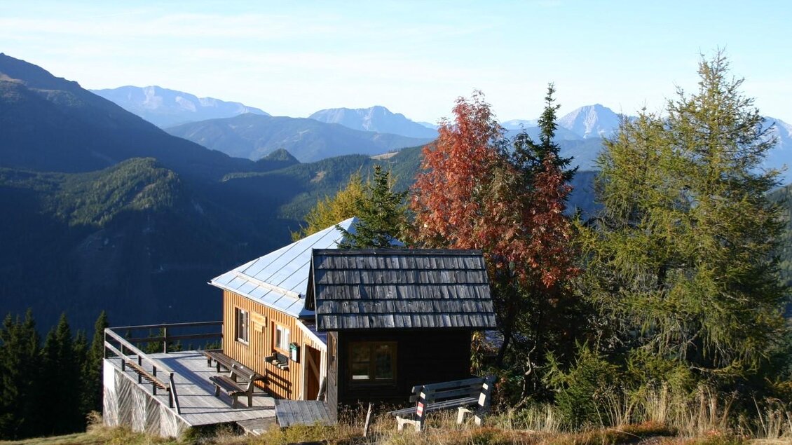 Eine gemütliche Hütte in den Bergen, umgeben von Bäumen und einem weiten Panorama. Der klare Himmel und die Berge im Hintergrund verleihen der Szene eine ruhige Atmosphäre. | © Heinz Kolland
