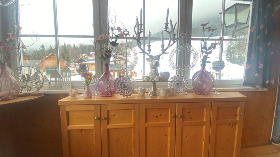 Eine Holzkommode mit verschiedenen dekorativen Vasen und Blumenarrangements. Im Hintergrund sind verschneite Berge und Häuser zu sehen. | © Bettina Scheck
