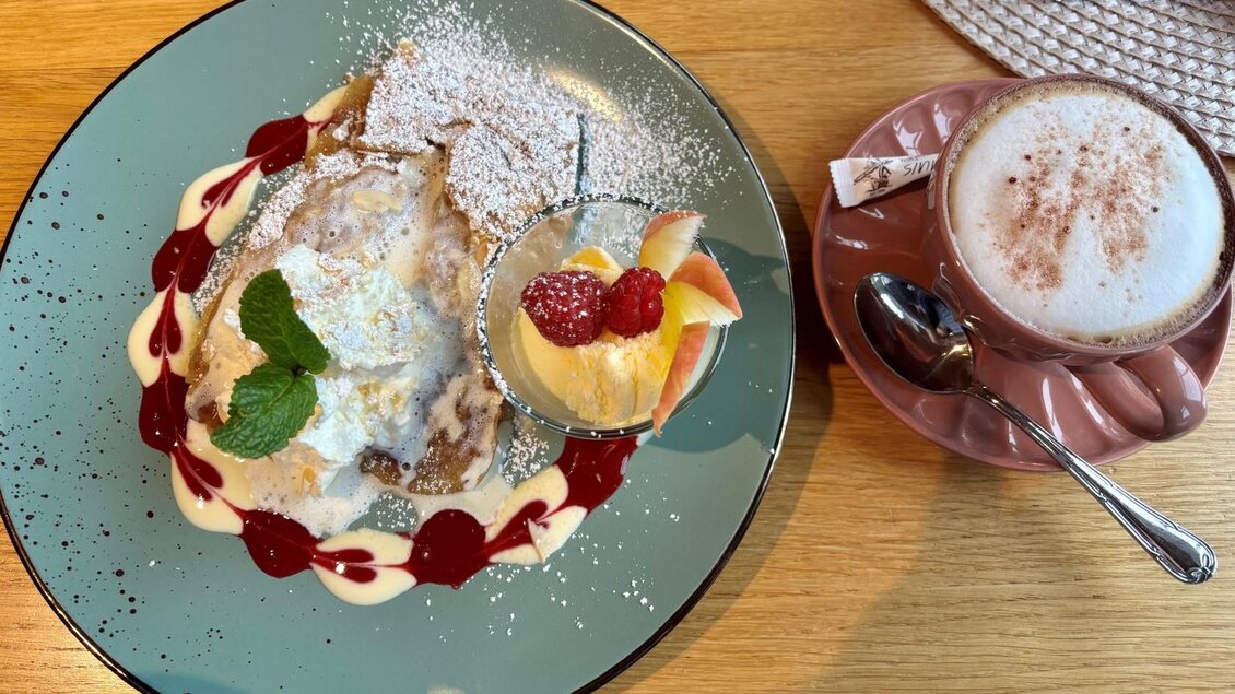 Ein leckerer Dessertteller mit Apfelstrudel, Sahne und Obst, garniert mit Puderzucker und Fruchtsauce. Daneben ein Becher heißer Schokolade. | © Hannah Scheck