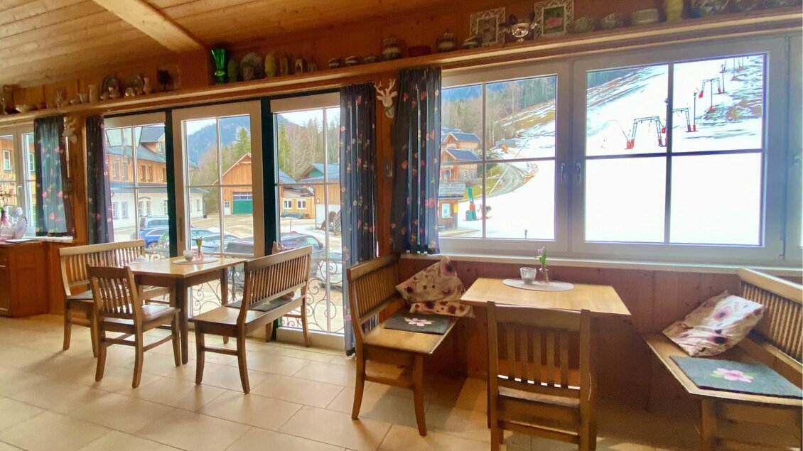 Ein gemütliches Café mit Holzinnenraum und großen Fenstern. Die Aussicht zeigt eine schneebedeckte Landschaft und nahe gelegene Hütten. | © Bettina Scheck