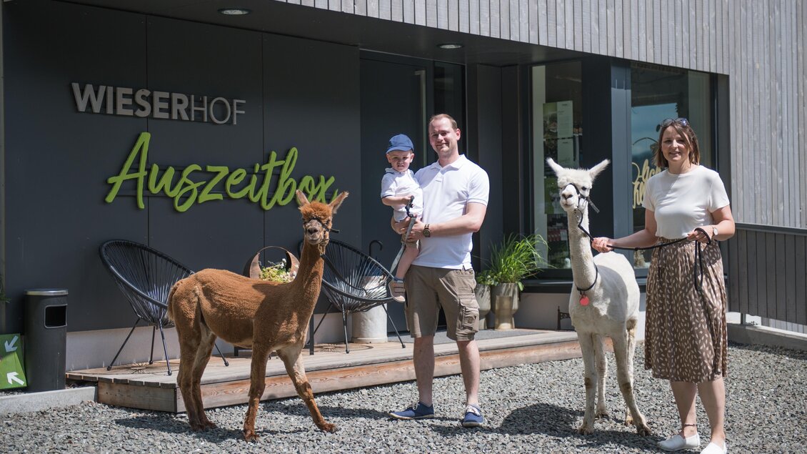 Eine Familie steht vor dem Wieserhof mit zwei Alpakas. Der Erwachsene hält ein Kind und lächelt in die Kamera. | © Anita Fössl