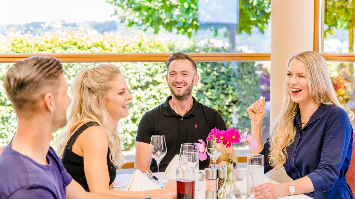 Eine fröhliche Gruppe von vier Personen sitzt an einem Tisch in einem hellen Restaurant. Sie genießen ein Gespräch und haben Getränke vor sich. | © Mias Photoart