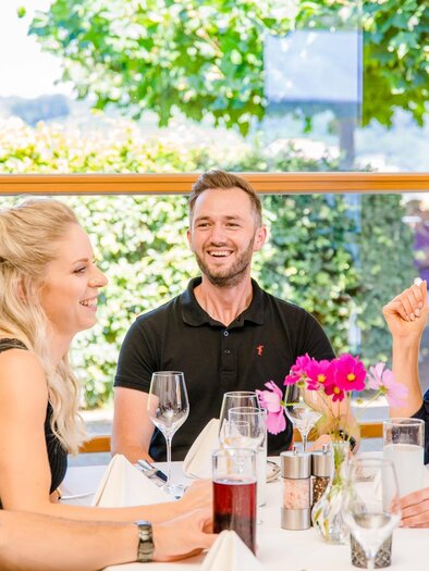 Eine fröhliche Gruppe von vier Personen sitzt an einem Tisch in einem hellen Restaurant. Sie genießen ein Gespräch und haben Getränke vor sich. | © Mias Photoart