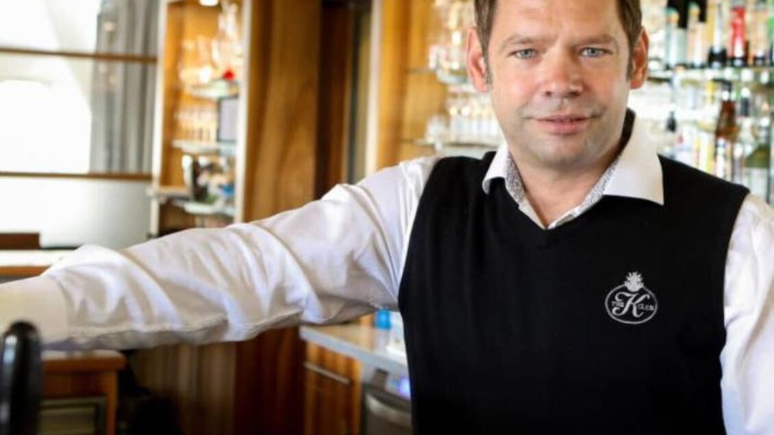 Ein freundlicher Barkeeper steht hinter der Theke in einer Bar. Im Hintergrund sind Regale mit Gläsern und Getränken zu sehen. | © Willibald Frauwallner