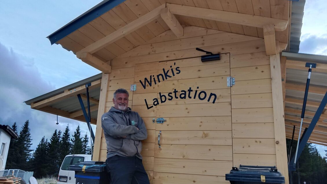 Ein Mann steht lässig vor einem hölzernen Gebäude mit der Aufschrift „Winki's Labstation“. Im Hintergrund sind einige Bäume und Fahrzeuge zu sehen. | © Winkelmayer