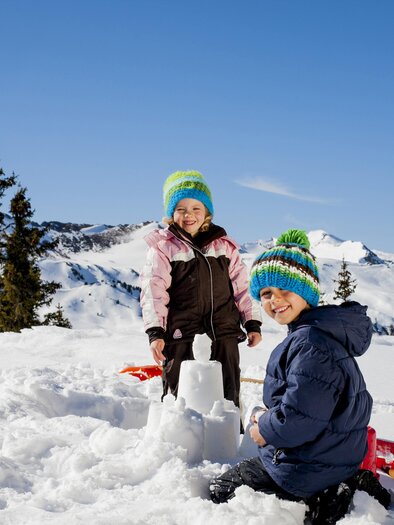 Two children are playing in the snow and building a snowman. In the background, there are snow-covered mountains and a blue sky. | © STG